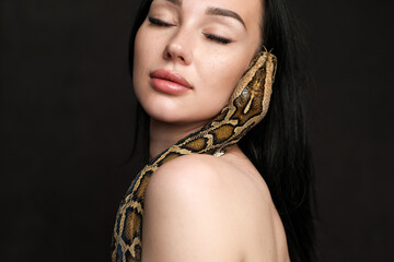 Portrait of a beautiful woman with dark hair on a dark background with a python near her face. Copy space