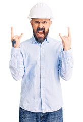 Young handsome man wearing architect hardhat shouting with crazy expression doing rock symbol with hands up. music star. heavy concept.