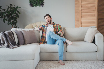 Full size photo of nice young man sit couch tired yawning watch tv wear stylish checkered shirt modern light cozy interior flat indoors