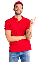 Young handsome hispanic man wearing delivery courier uniform smiling happy pointing with hand and finger to the side