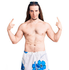Fototapeta premium Young adult man with long hair wearing swimwear shirtless looking confident with smile on face, pointing oneself with fingers proud and happy.