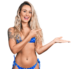 Young blonde woman wearing bikini amazed and smiling to the camera while presenting with hand and pointing with finger.