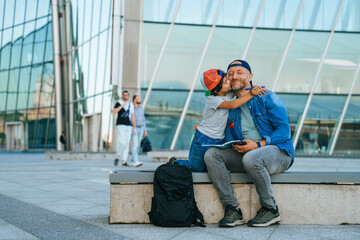Father and son hugging on a bench outdoors, child giving a kiss, playful moment near modern building