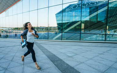 Professional woman running with folders in hand, outdoors near modern glass building, rushing to an appointment