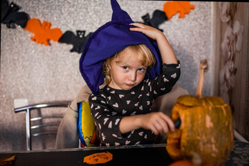 A child in a witch's hat carves a pumpkin for Halloween in a decorated indoor space