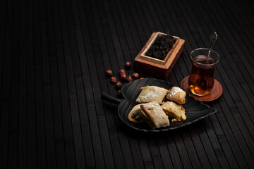 Minimalistic scene with traditional pastries on a black plate, hazelnuts, and a glass of tea. A rustic wooden box with loose tea leaves enhances the cozy, authentic tea-time atmosphere.