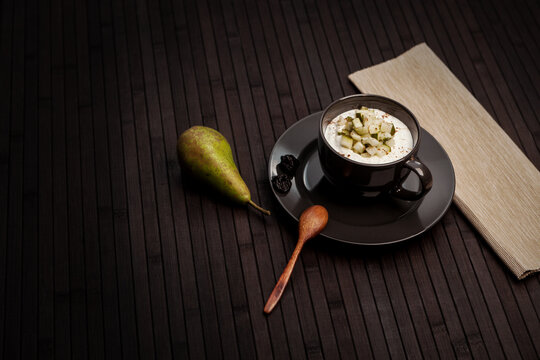 A dark, minimalistic setup featuring a black cup filled with creamy dessert topped with diced pear. A whole pear, dried prunes, and a wooden spoon complete the rustic, cozy atmosphere.