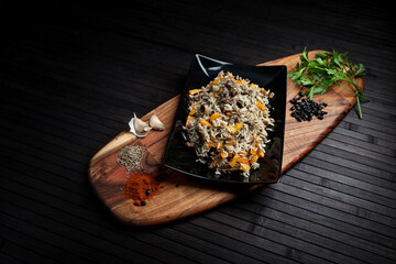 A rustic plate of rice pilaf with carrots and meat, garnished with spices. Presented on a wooden board with garlic, cumin, parsley, and dried berries, creating a hearty and traditional meal setting.