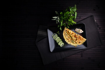 A beautifully plated omelette garnished with herbs, served with cucumber slices and a side of dip on a sleek black plate. Fresh herbs complete the minimalistic, elegant dining setup.
