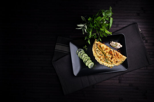 A beautifully plated omelette garnished with herbs, served with cucumber slices and a side of dip on a sleek black plate. Fresh herbs complete the minimalistic, elegant dining setup.