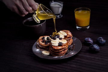 A stack of pancakes topped with banana slices and berries, being drizzled with honey. A cup of coffee, a glass of juice, and plums complete the cozy breakfast setup.