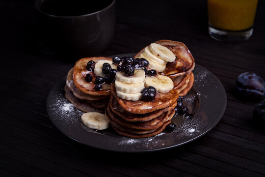 A close-up of a stack of pancakes topped with banana slices and blueberries, dusted with powdered sugar. A cup of coffee, a glass of juice, and fresh plums complete the cozy breakfast setting.