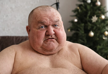 Obraz premium Overweight man, 50-60, sweating in warm winter temperatures, sitting by Christmas tree, single and divorced, reflecting on life during holidays.