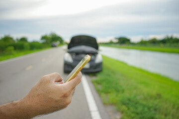Close-up of male driver's hand holding a smartphone calling mechanic with a broken car on the side...
