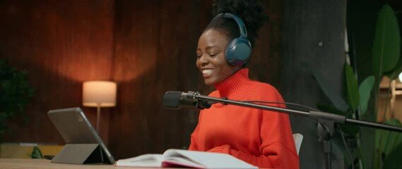 Horizontal footage of smiling beautiful young African American woman with headphones sitting at workplace. Female radio host using microphone during working day. Girl using digital tablet. Parallax