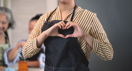 A woman wearing an apron and holding a heart in her hand. She is smiling and surrounded by other people
