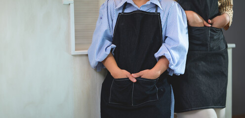 Two women wearing aprons and standing next to each other. One of them is holding her hand in her pocket