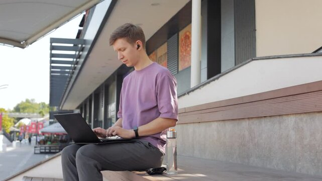 A young man listening to music with wireless headphones and using a laptop outside. Writing on a computer.