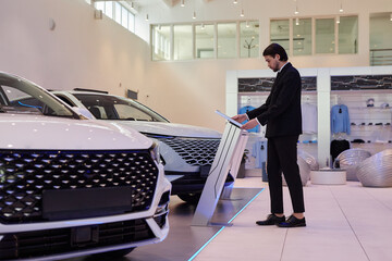 Side view of Central Asian male auto dealer in formal black suit checking vehicle spec sheets displayed on information stand at car dealership center, copy space