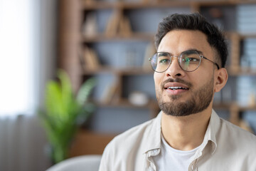 Fototapeta premium Hispanic man with glasses at home looking content and thoughtful in modern indoor setting