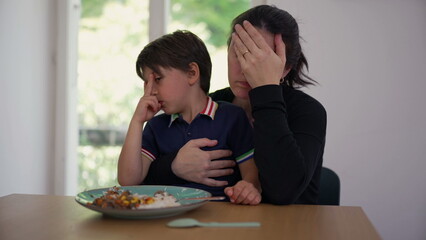 Mother and older son rest their heads on the dining table, looking tired. The moment captures a shared sense of fatigue and connection between them, highlighting the realities of family life