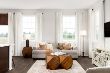 A cozy living room detail with a wooden table on an area rug and hardwood floors, orange and cream pillows sit on a tan couch, and white curtains are hung aside the windows. No names or labels.