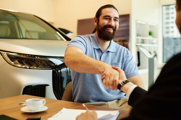 Medium shot of happy adult man shaking hands with auto dealer after successful car buying and...