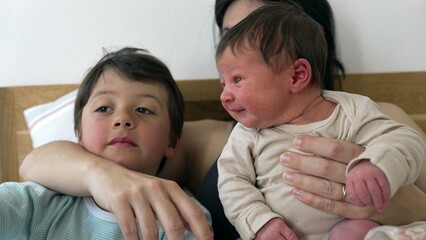 Older sibling and mother hold baby together, with sibling looking at the baby curiously. The moment captures the bond and affection shared within the family, highlighting love and connection