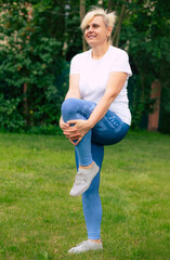 Smiling woman in sportswear performs a stretching exercise, pulling one knee to the chest while balancing on one leg in a grassy outdoor. The image suggests warm-up, flexibility, and fitness focus.