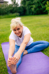Fototapeta premium A smiling female performs a seated stretch on a purple yoga mat in a grassy outdoor area, reaching for their foot. The image conveys flexibility, well-being, and a joyful fitness routine in nature.