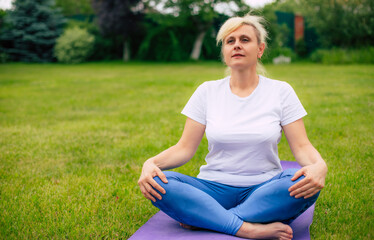 Sporty adult woman sits cross-legged on a purple yoga mat, meditating in a peaceful outdoor setting. The image highlights mindfulness, relaxation, and the calmness of practicing meditation in nature