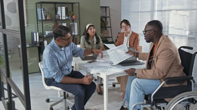 Full slowmo shot of African American businessman in wheelchair brainstorming ideas for joint project with multiracial business partners at office meeting, inclusion and accessibility