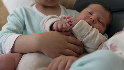 Older sibling holds baby while both lie on the bed. The baby looks up curiously, showcasing a tender moment of sibling bonding and family love in a cozy and intimate home setting