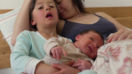 Older sibling gently cradles baby while resting on mother’s lap. The siblings share a peaceful moment, highlighting the close bond and affection between them in a cozy home environment
