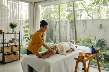 Woman providing soothing leg massage to client lying on massage table in calm, light-filled room...