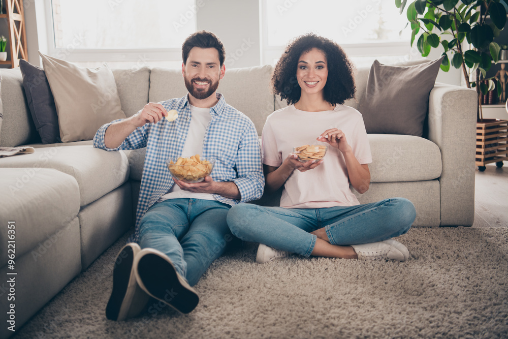 Poster full size photo of two nice young partners eat chips sit floor weekend modern apartment indoors