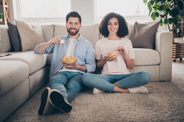 Full size photo of two nice young partners eat chips sit floor weekend modern apartment indoors