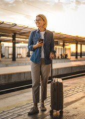 Mature woman wait for the train and hold cellphone and suitcase