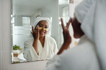 African American woman applying under-eye patches while looking in bathroom mirror, wearing bathrobe and towel on head. Reflecting in large, bright bathroom mirror while performing skincare routine