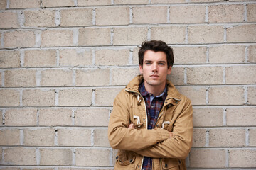 Obraz premium Arms crossed, portrait and man with fashion by brick wall downtown for holiday and vacation for city culture. Serious, jacket or student with trendy winter clothes and cool streetwear style in Spain