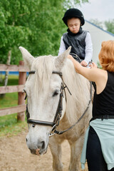 Child riding white horse with guidance from trainer in outdoor setting. Equestrian training session taking place near wooden fence and green trees on clear day