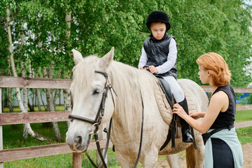 Boy receiving riding lessons on a white horse with instructor's assistance. Scene set outdoors with trees and wooden fence as background