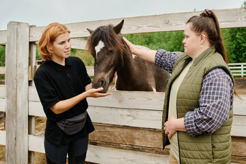Two women interacting with horse, feeding it in rural barn setting. Both women showing gentle...