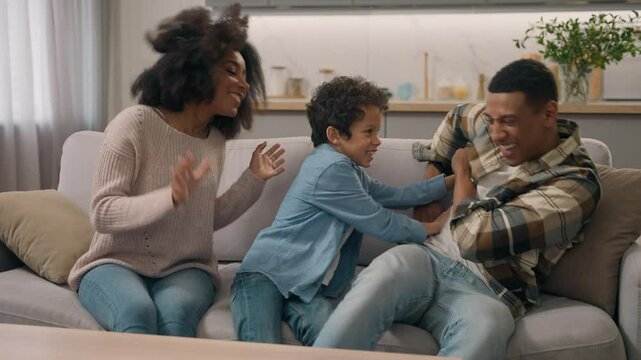 Happy family having fun African American joyful couple female mom woman male dad man cuddle hug embrace playing tickling their adopted child little boy laughing son kid inside modern living room flat