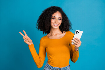 Photo portrait of attractive young woman hold device show v-sign dressed stylish yellow clothes isolated on blue color background