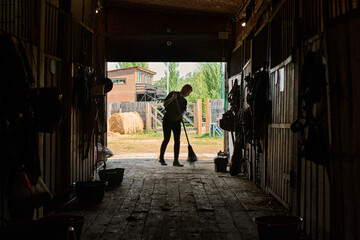 Person in silhouette cleaning barn floor with a broom, surrounded by equipment and horse stalls, sunlight streaming through open barn door leading outside