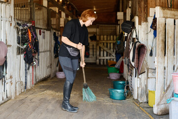 Person cleaning stable corridor with a broom while standing amid various equipment hung on wooden walls creating an organized and neat workspace