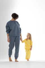 Fototapeta premium Young mother and little daughter walking holding hands. Dressed in yellow and ment color suits on white background, studio. Concept of advertising positive and clothes. High quality photo