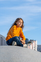 A young girl wearing a bright orange sweater sits calmly on a large rock, enjoying the sunny day and city backdrop