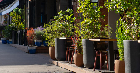 A restaurant features an inviting outdoor seating area lined with vibrant green plants and flowers on a bright, sunny day
